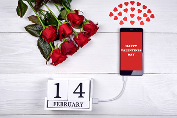 Valentines Day background: bouquet of red roses, cellphone with sign Happy Valentines Day charging with february 14 wooden block calendar, copy space. Greeting card mockup