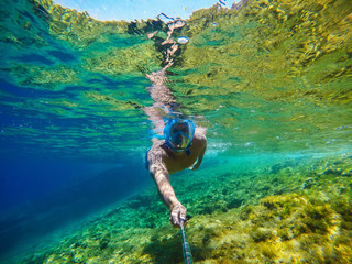 Underwater photo of a young tourist man swimming in the turquoise sea under the surface with snorkelling mask for summer vacation while taking a selfie with a stick.