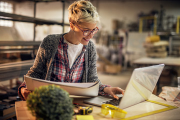 Close up view of charming smiling motivated short hair attractive middle aged industrial female engineer with eyeglasses working with blueprints and laptop in the workshop.