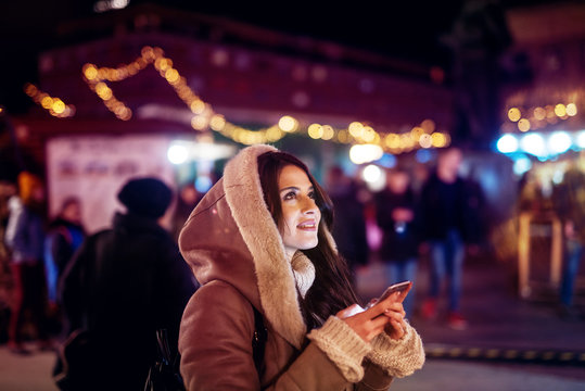 Close Up Side View Of Hooded Satisfied Cheerful Stylish Attractive Beautiful Young Happy Girl In Sweater And Jacket Looking At The Sky While Holding A Mobile On The Night City Street.