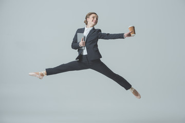 businesswoman in suit and ballet shoes jumping with coffee and tablet, isolated on grey