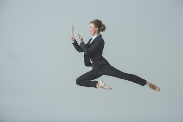 businesswoman in suit and ballet shoes jumping and using digital tablet, isolated on grey