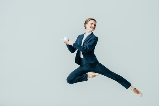 businesswoman in suit and ballet shoes jumping with cup of coffee, isolated on grey