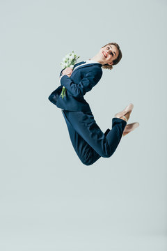 businesswoman in suit and ballet shoes jumping with bouquet of flowers, isolated on grey