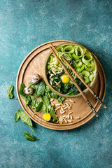 Ceramic bowl with vegetarian green pea noodles, sliced cucumber, celery, spinach, quail egg yolk, pine nuts with wooden chopsticks. Turquoise texture background. Top view, space. Healthy eating.