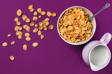 Bowl with corn flakes, jug of milk and spoon on purple background, top view
