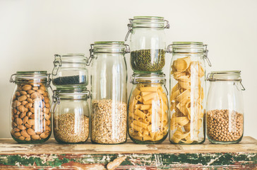 Various uncooked cereals, grains, beans and pasta for healthy cooking in glass jars on wooden table, white background, horizontal composition. Clean eating, vegan, balanced dieting food concept