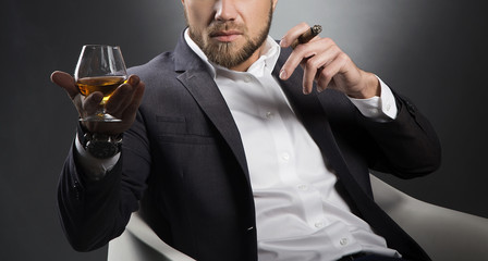Cognac glass and cigar in hands of male model wearing suit. Man with beard with brandy smoking