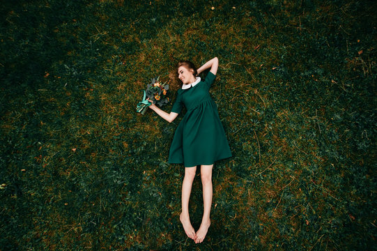 Young Girl With Red Hairlying On Ground. Portrait From Above Of Cute Teenager Female Relaxing On Grass In Summer. Lovely Skinny Female In Beautiful Stylish Dress Holding Bouquet Of Flowers In Hand.