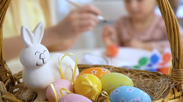 Happy Easter! Young mother and her cute little daughter wearing funny rabbit ears are coloring easter eggs sitting at a festive table with basket and Bunny. Slow Motion