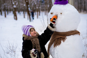 Fototapeta na wymiar A small cheerful girl holds a big carrot, the nose of a big snowman. A cute little girl has fun in winter park, wintertime