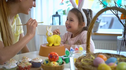 Happy Easter! Mother and her little daughter with bunny ears painting Easter Bunny at the festive table with basket and eggs