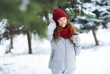 Fototapeta na wymiar Outdoor close up portrait of young beautiful girl with hot cup of coffee. Girl dressed in warm winter clothes and a hat posing in a winter forest. Christmas, winter holidays concept. 