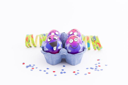 Group of colorful painted Easter eggs with funny cartoon style faces in a light blue egg box, colorful confetti and paper streamer on white background