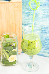 Making Tropical Healthy Smoothie Concept. A glass of green smoothie and a jar of cut fruit and spinach against a light colored wooden wall