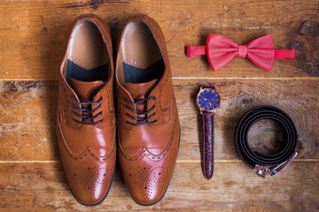 Closeup of elegant stylish brown male formal accessories isolated on brown wooden background. Top view of leather belt, shoes, watch and bowtie. Preparation for wedding or bussiness day concept. 