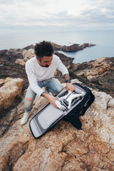 Fototapeta premium Professional stock aerial photographer, urban adventurer and location explorer, traveler unpacks backpack with expensive equipment and drone, ready to fly over oceanside and inspiring scenery