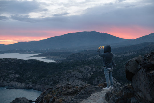 Unrecognizable man in hoodie stands on top of hiking trail on mountain, overlooks amazing beautiful scenery with sunset, makes photo on smartphone camera, concept lifestyle travel blogger