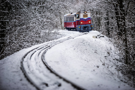 Fototapeta Budapest, Hungary - Beautiful winter forest scene with snow and old colorful train on the track in the Hungarian woods of Huvosvolgy