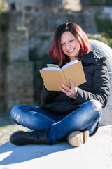 Obraz premium Happy smilling handsome redhead young woman sitting on a small wall and reading a book - Concept Relaxing in nature.