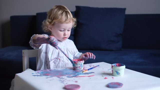 Toddler girl painting with paintbrush and colorful paints on white paper at the table
