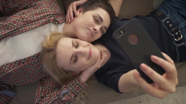 Two young girls lie on the couch, do selfie on a smartphone, smiling, lovers, lgbt, young couple, top shot 60 fps