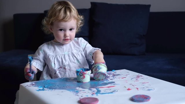 Toddler girl painting with her hands using colorful paints on white paper at the table