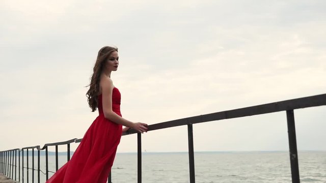a young woman in a long scarlet dress holds onto the railing near the sea, her hair and hem of a red evening dress fluttering under the streams of wind near the sea