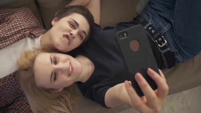 Two young lesbian girls lie on the couch, do selfie on a smartphone, lovers, lgbt, young couple, teenagers top shot 60 fps