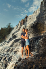 Obraz premium a young couple is standing in an embrace at the foot of a waterfall, seeing off the passing sun