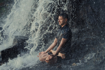 a man sits under a waterfall in a lotus pose and meditates, his eyes are closed