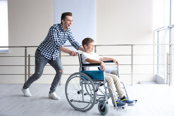 Young man having fun with little boy in wheelchair indoors