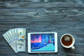 Tablet computer with stock data, cup of coffee and money on wooden background, top view