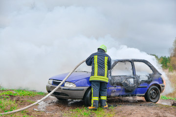 Plakat Fire fighter prepare to attack a propane fire. Burning and crashed car after explosion. Accident on street at countryside. No one was injured. Artificially created set for film making