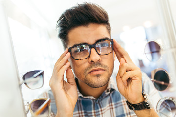 Handsome young man choosing eyeglasses frame in optical store.