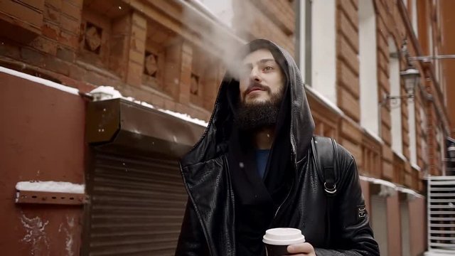 Stylish man with a beard dressed in black leather clothes is going day through the old town in the snow and smoke a cigarette while drinking morning coffee