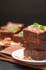 Chocolate brownie square pieces in stack on white plate with walnuts, decorated with mint leaves and cocoa on black background. Delicious dessert. Dark mood. Close up photography. Selective focus