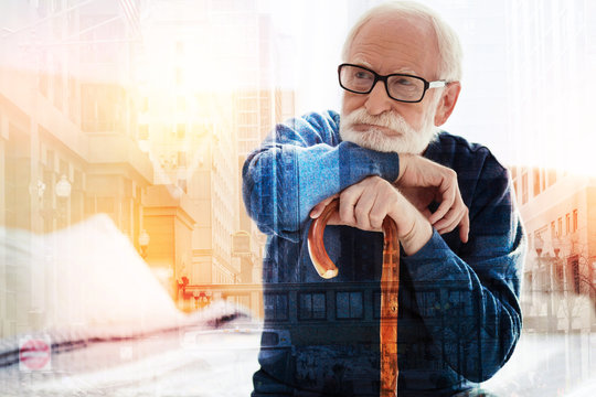 Thoughtful glance. Calm wise aged man sitting with a walking stick in his hands and thoughtfully looking into the distance
