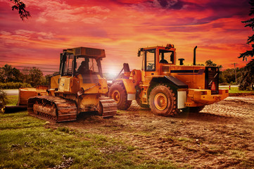Large bulldozers at construction site, cloudy sky and sunset in background.