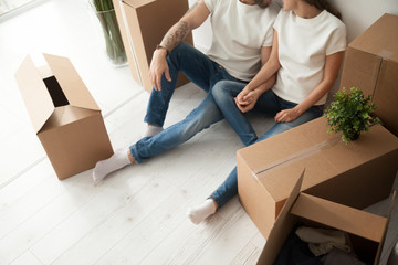 Close up view of happy couple sitting on the floor with cardboard boxes holding hands ready to relocate move in out house, young man and woman packed belongings on moving day settling in new own home