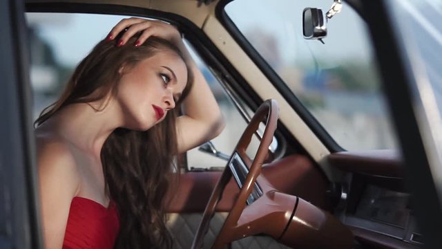 a young and beautiful woman with long curly hair is dressed in a red dress, the lady is inside the vehicle, she put her hands on the steering wheel and looks in the mirror