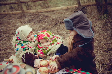 The children in an old fur coat and ornament scarf eating cheesecake at the holiday table. Shrovetide Maslenitsa