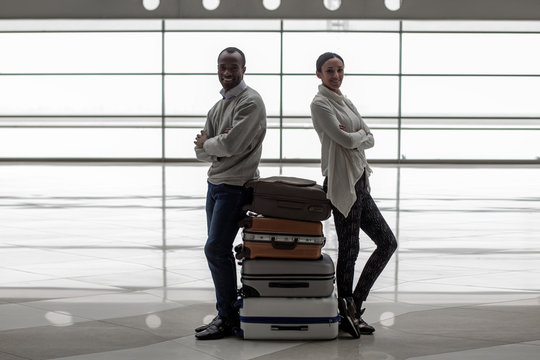 Honeymoon. Full length portrait of happy young enamored man and woman are leaning on suitcases with crossed arms at airport lounge. They are standing back to back and looking at camera with joy
