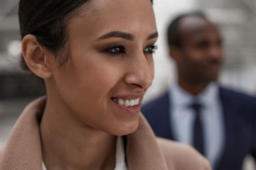 Confident glance. Close-up of face of gorgeous charming young woman is looking aside with smile. She is standing with african man in background. Selective focus