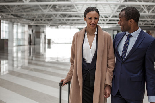Travel abroad. Portrait of gorgeous young charming lady is standing with african guy in airport hall. She is looking at camera with smile while holding handle of her luggage. Copy space in left side