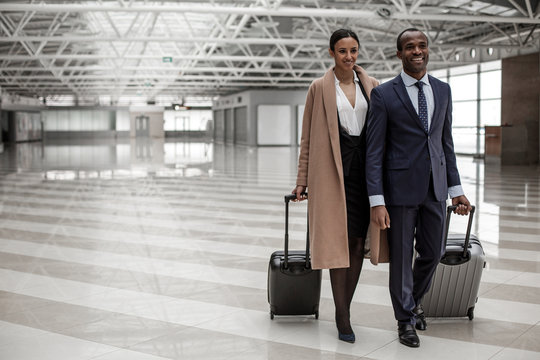 Fool of joy. Full length portrait of happy elegant business partners are walking along airport lounge while carrying luggage. They are looking ahead with smile. Copy space in the left side