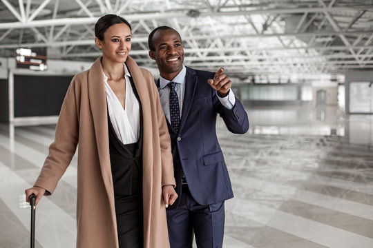 Look at that. Portrait of cheerful young man and woman are standing at terminal lounge with suitcases. Excited businessman is pointing finger forward while female is smiling. Copy space in right side