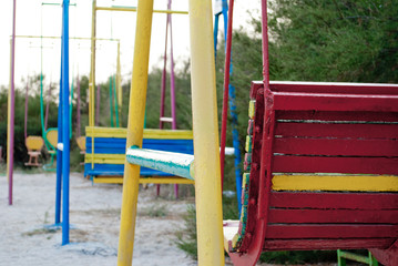Fototapeta na wymiar Multicolored childrens swing from metal, high, against a background of green tree bush, on sand summer sun relaxing, playground, blue sky