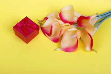 Fototapeta premium Bouquet of red yellow calla lilies with red gift box and ribbon on yellow cardboard background.