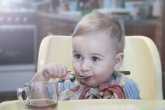 Cute little boy drinking tea from a mug with a spoon while sitting in a chair